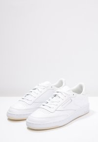 Ett par vita låga sneakers med snörning, perforerat tåområde och subtila sömnadsdetaljer på en neutral vit yta.