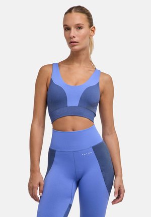Giovane donna che indossa un reggiseno sportivo blu senza cuciture e leggings a vita alta con pannelli laterali patternati, in piedi su uno sfondo semplice.