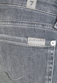 Vaqueros de mezclilla gris con una etiqueta que dice "para toda la humanidad". Los detalles notables incluyen patrones de costura y un botón metálico como acento.
