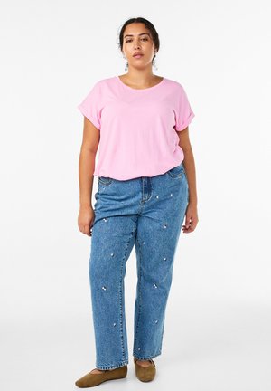 Žena stojaca oblečená v ružovej tričku s krátkym rukávom, modrých džínsach s malými vyšívanými kvetmi a hnedých plochých topánkach na bielom pozadí.