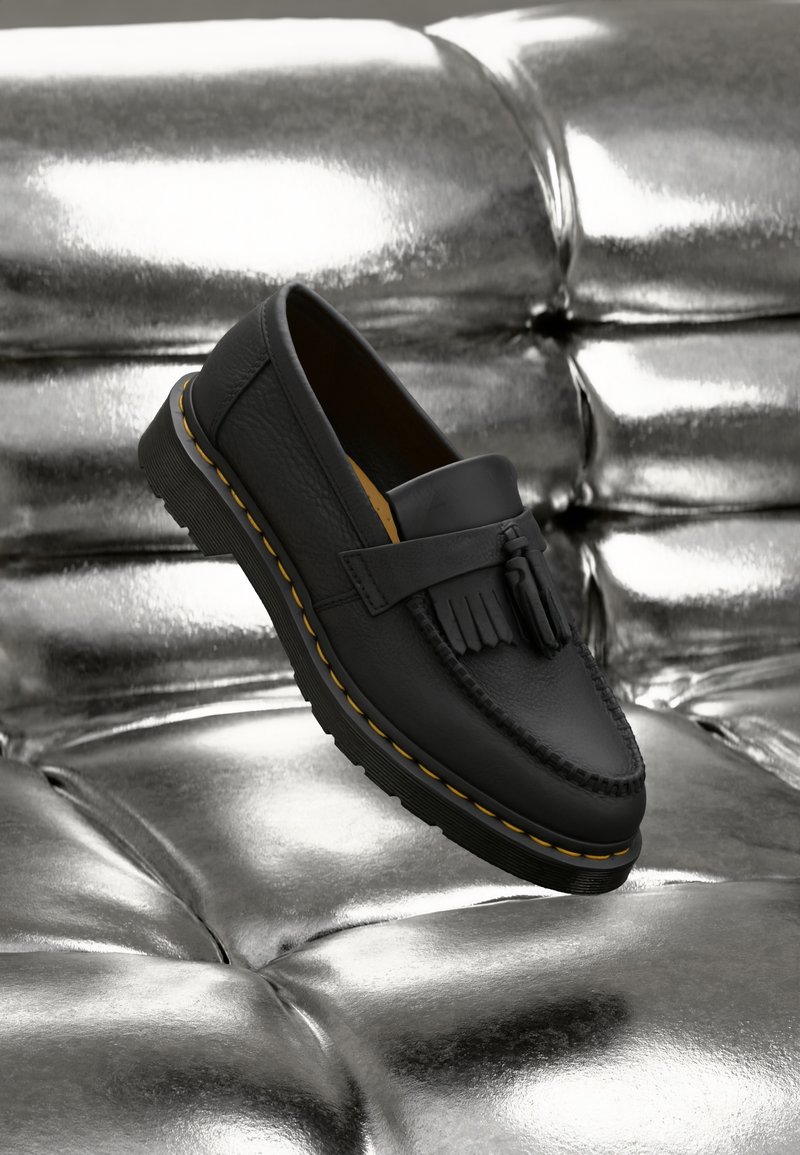 Mocassin en cuir noir avec pompons et coutures jaunes sur une semelle épaisse, posé sur une surface argentée tuftée brillante.
