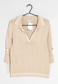 Maglione lavorato a maglia beige con un ampio colletto, zip frontale a metà e dettagli a strisce. Presenta maniche a tre quarti con cordini regolabili.