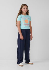 Helles, blaues Baumwoll-T-Shirt mit orangefarbener Grafik, Text und Sonnenmotiv, kombiniert mit marineblauen, weiten Hosen. Weiße Sneaker mit sandfarbenen Sohlen.