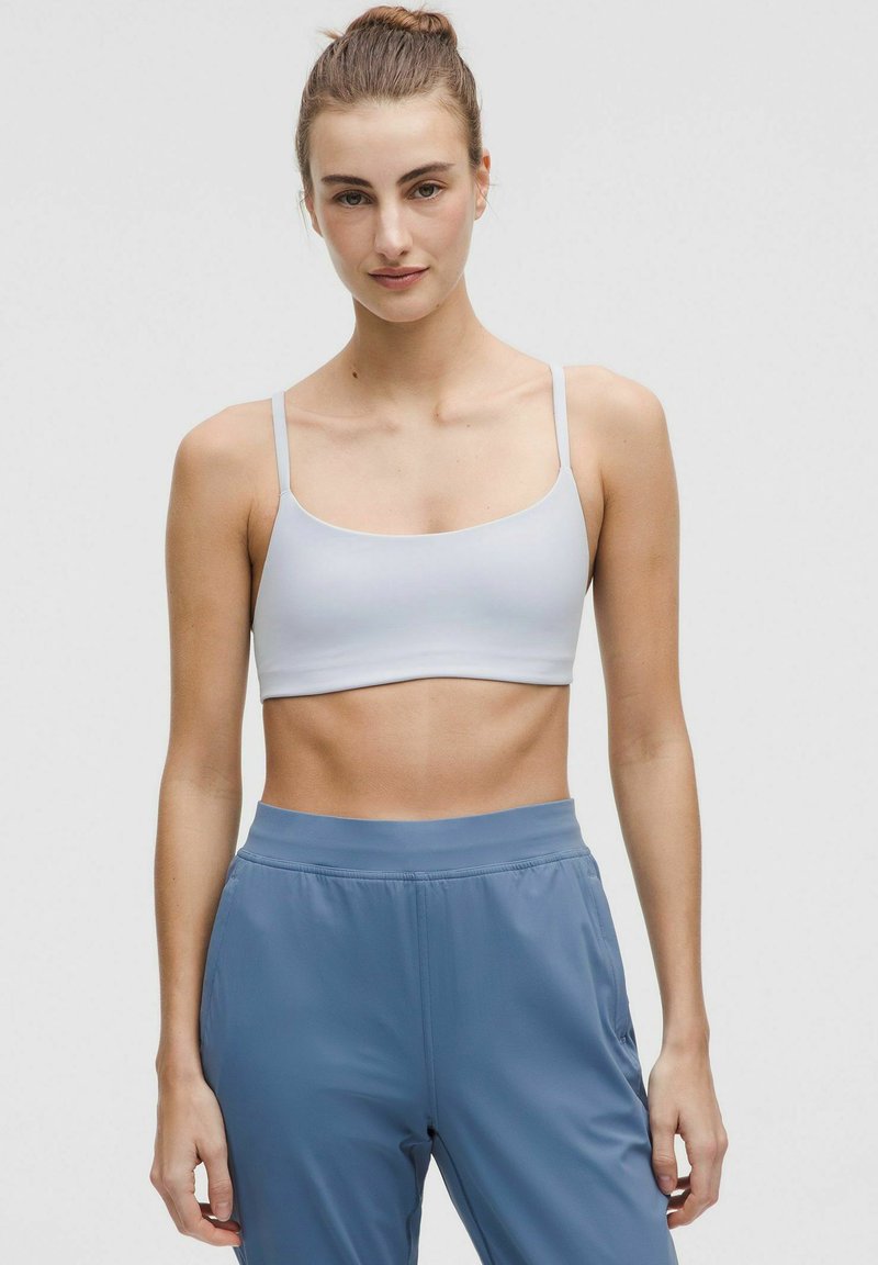 Sujeción deportiva gris claro con tirantes finos y escote redondeado, combinado con pantalones deportivos azules que cuentan con una cinturilla elástica.