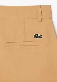 Beige kalhoty s hladkým povrchem, úzkým střihem a malým zeleným vyšitým krokodýlem na zadní kapse.