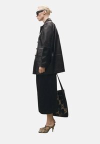 Black leather jacket with front buttons, black midi skirt, snake print mules, and a patterned black tote bag. Long, straight blonde hair.
