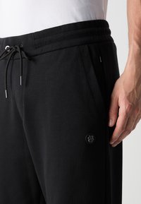 Čierne teplákové nohavice z mäkkého materiálu s elastickým pásom a šnúrkou. Obsahujú bočné vrecká a čiernu logo nášivku na stehne.