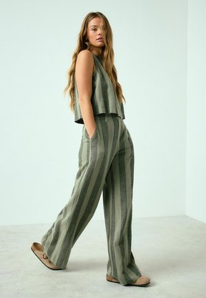 Young woman walking indoors wearing green striped sleeveless top and wide-leg pants with beige slip-on shoes.