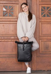 Sac à dos noir à rabat en cuir texturé, avec fermeture à boucle, reposant sur une tenue gris clair sur fond en bois.