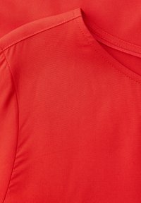 Gros plan sur un tissu en coton rouge montrant l'encolure et la couture d'épaule avec des détails de surpiqûres visibles.