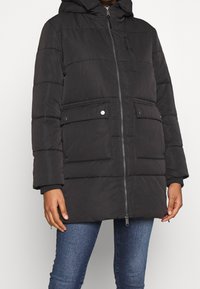 Osoba v černé prošívané zimní bundě s kapucí, předním zipem, kapsami se zvoněnými knoflíky a modrých džínách na bílém pozadí.