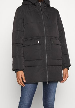 Osoba w czarnej pikowanej, zimowej kurtce z kapturem, z zamkiem błyskawicznym z przodu, kieszeniami z klapami na zatrzaski i niebieskich dżinsach na białym tle.