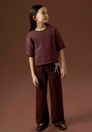 Maglione a maglia color bordeaux con maniche corte e motivo testurizzato, abbinato a pantaloni larghi bordeaux dotati di un nodo decorativo e scarpe scure.