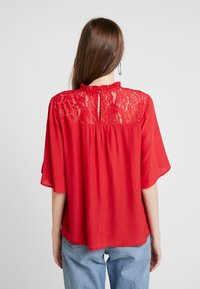 Rød bluse med blondedetalje på ryggen, korte flared ærmer, høj halsudskæring med flæser og en nøglehulldetalje på ryggen. Blødt stoftekstil.