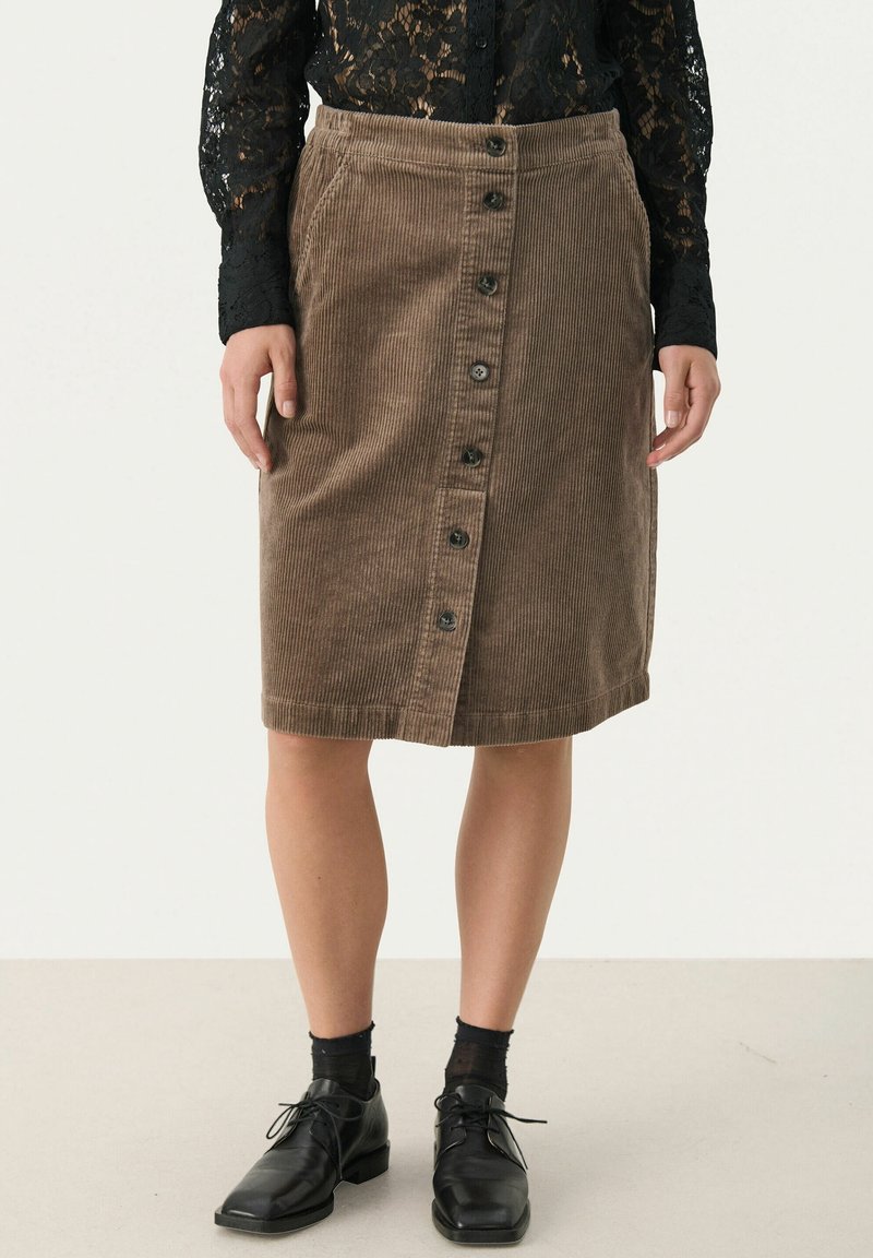 Femme portant une jupe en velours côtelé marron à boutons sur le devant, longueur genoux, un haut en dentelle noire à manches longues, des socquettes noires et des chaussures noires à lacets.