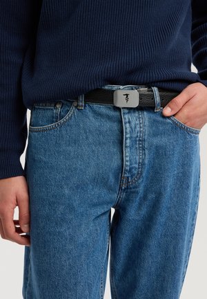 Cinturón de cuero negro con hebilla metálica gris, con un logo; lleva sobre unos vaqueros azules y un suéter de punto oscuro.