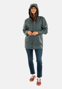 Waterdichte groene parka jas met een capuchon, magnetische sluiting aan de voorkant, twee zijzakken en verstelbare trekkoorden. Draagt over jeans en sneakers.