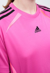 Detailný záber na osobu oblečenú v jasne ružovom športovom tričku Adidas s čiernymi pruhmi na pleciach a logom na hrudi.