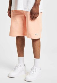 Shorts de couleur pêche en tissu doux, dotés d'une taille élastique et d'un détail logo blanc. Assortis à des baskets blanches et des chaussettes.