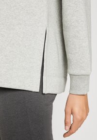 Jasnoszara bluza z bocznymi rozcięciami, długimi rękawami, ściągaczami przy mankietach i miękką fakturą, zestawiona z ciemnoszarymi legginsami.