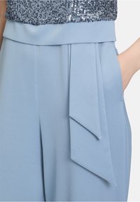 Pantalons taille haute bleu clair avec une texture lisse, dotés d'un accent de nœud à l'avant et de poches latérales, complétés par des plis.