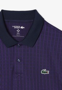 Lila polo skjorta med rutmönster, marinblå krage, tre knappar och Lacoste-logotyp på vänster bröst. Slim fit-design och texturerad tyg.
