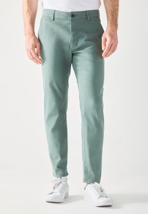 Green cotton pants with a slim fit, front pockets, and a single button closure. Paired with white sneakers and a plain white t-shirt.