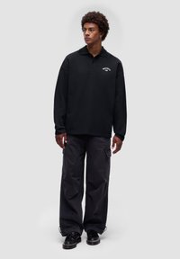 Schwarzes Langarm-Polohemd mit kragenförmigem Halsauschnitt und gesticktem Logo, kombiniert mit locker sitzenden dunklen Hosen und schwarzen Schuhen.