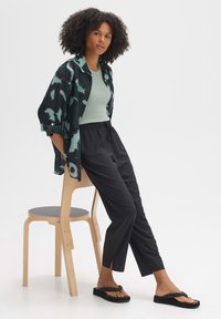 Schwarz-türkis gemustertes Hemd, grüner Tanktop, schwarze tapered Pants und schwarze Flip-Flops, vor einem neutralen Hintergrund mit einem Stuhl.