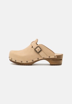Beige leren klomp met houten zool, verstelbare gesp en metalen studs langs de rand van de zool, getoond op een effen achtergrond.