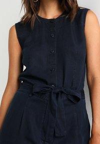 Tmavě modrý bezramenní overal s předním zapínáním na knoflíky, uvázaným páskem v pase a hladkou texturou látky. Disponuje strukturovaným výstřihem a bočními kapsami.