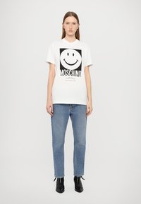 Bílý bavlněný tričko s černým grafickým potiskem usměvavého obličeje a textem "MOSCHINO", doplněné modrými džíny a černými kotníkovými botami.