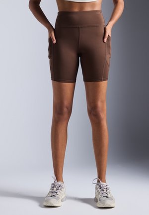 Femme portant un short de cyclisme marron taille haute avec des poches latérales et des baskets blanches, debout devant un fond neutre.
