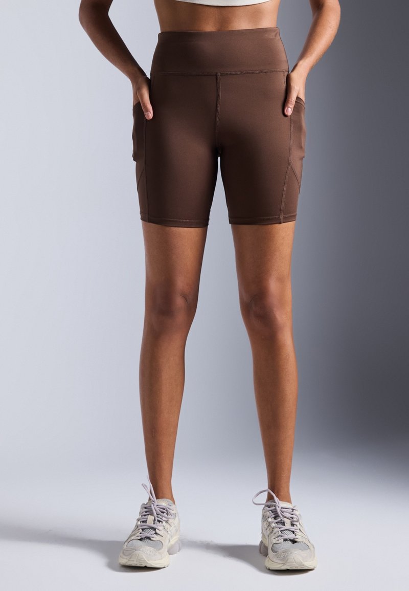 Femme portant un short de cyclisme marron taille haute avec des poches latérales et des baskets blanches, debout devant un fond neutre.