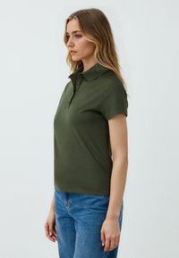 Polo shirt verde oliva, maniche corte, con bottoni sulla parte anteriore, realizzato in tessuto morbido. Abbinato a jeans azzurri.