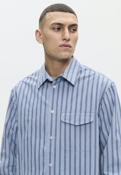 Joven con el pelo corto que lleva una camisa de botones azul y blanca con rayas verticales y un bolsillo en el pecho, mirando hacia un lado contra un fondo liso.