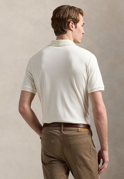 Kremfarget polotrøye med korte ermer og krage, kombinert med brune bukser. Skjorten har en subtil tekstur og kontrasterende kant på kragen.