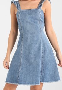 Denimmekko, jossa on neliön muotoinen kaula-aukko, rypytetyt hihnat ja liehuva hame. Vaalea sininen väri, jossa on revitty helma ja selkeät saumat.