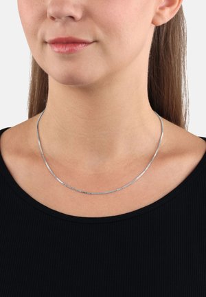 Donna che indossa una sottile collana d'argento sopra un top nero, mostrando il collo e la parte bassa del viso con le labbra chiuse e i capelli lisci.