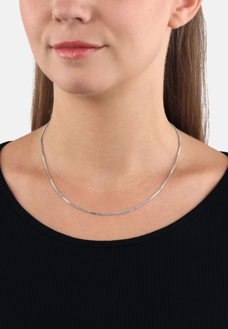 Donna che indossa una sottile collana d'argento sopra un top nero, mostrando il collo e la parte bassa del viso con le labbra chiuse e i capelli lisci.
