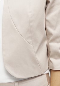 Détail du manche et de la couture latérale d'un blazer beige clair sur mesure porté par-dessus un vêtement blanc, gros plan montrant la texture du tissu et les coutures.