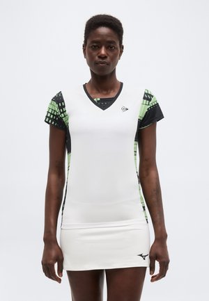 Mujer con vestido deportivo blanco con mangas cortas estampadas en verde y negro, de pie contra un fondo liso y claro, mirando hacia adelante.