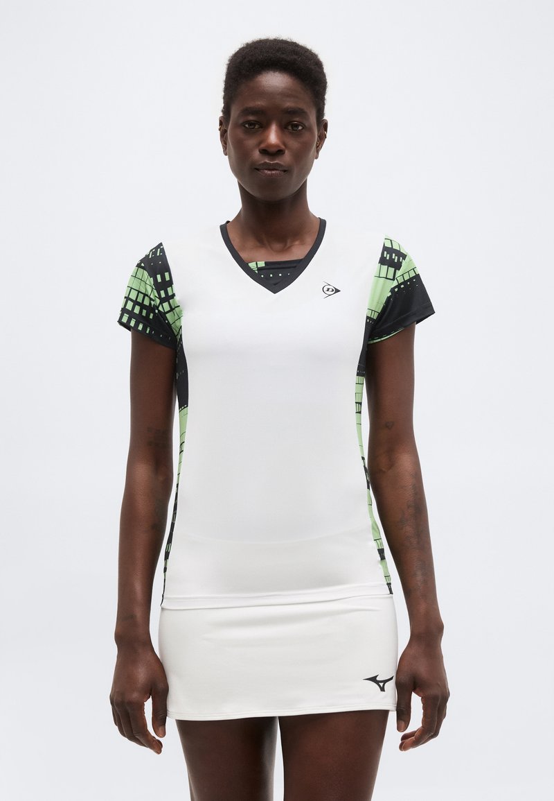 Femeie îmbrăcată într-o rochie sport albă cu mâneci scurte cu model verde și negru, stând pe un fundal simplu deschis, cu fața spre înainte.