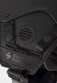 Tums melns ķivere tuvplānā ar teksturētu virsmu, regulējamiem siksnām, apaļu ventilāciju un logo uzlīmi ar uzrakstu "Sweet Protection."
