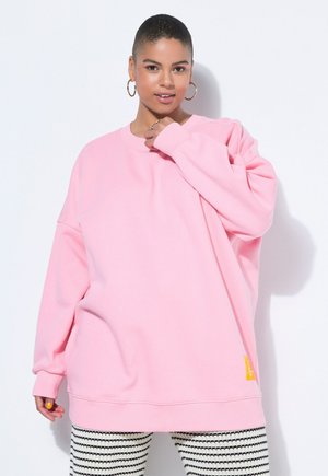 Femme aux cheveux courts portant de grandes boucles d'oreilles et un sweat-shirt rose ample, posant devant un fond clair.