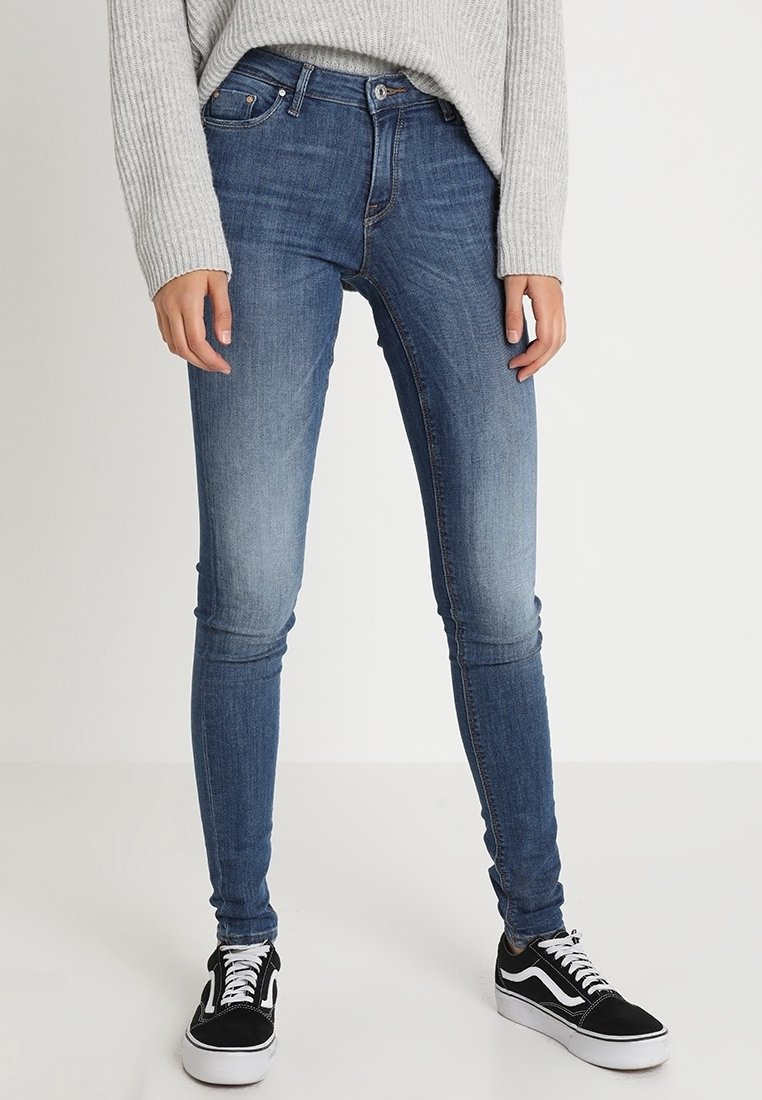 Personne portant un jean skinny bleu, des baskets noires et blanches, et un pull côtelé gris clair, debout devant un fond uni.
