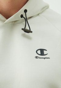 Svetlo šedá mikina s kapucňou vyrobená z mäkkého materiálu, ktorá obsahuje šnúrku s čiernymi detailmi a čierny logo "Champion" na hrudi.