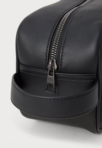 Sac en cuir noir avec une surface texturée, équipé d'une fermeture éclair proéminente et d'une poignée ronde. Design minimaliste avec des lignes épurées et du matériel sombre.