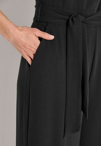 Hand in de zijzak van een zwarte broek met een vastgebonden stoffen riem om de taille.