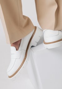 Białe skórzane mokasyny z grubą, solidną podeszwą i brązowymi przeszyciami. W zestawieniu z jasnobeżowymi spodniami, które charakteryzują się minimalistycznym stylem.